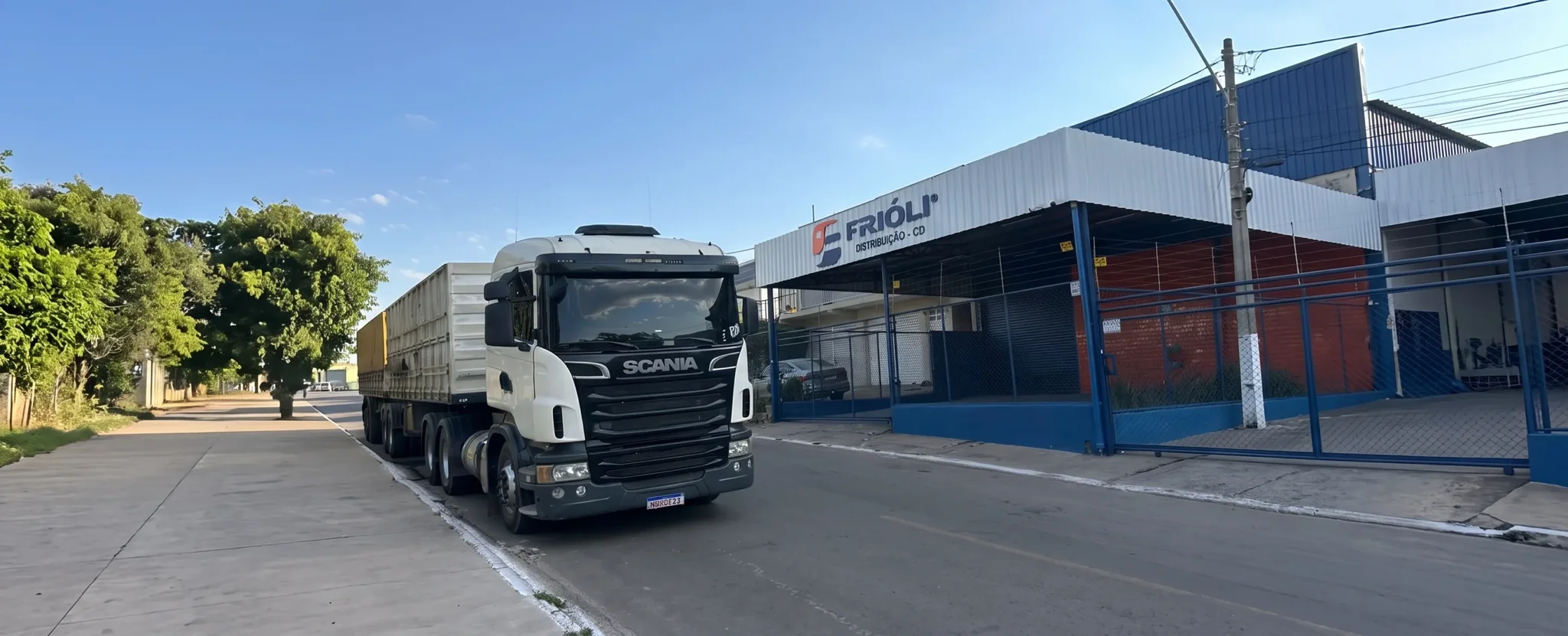 White Scania semi-truck parked outside the Frioli Distribuição building on a sunny day.